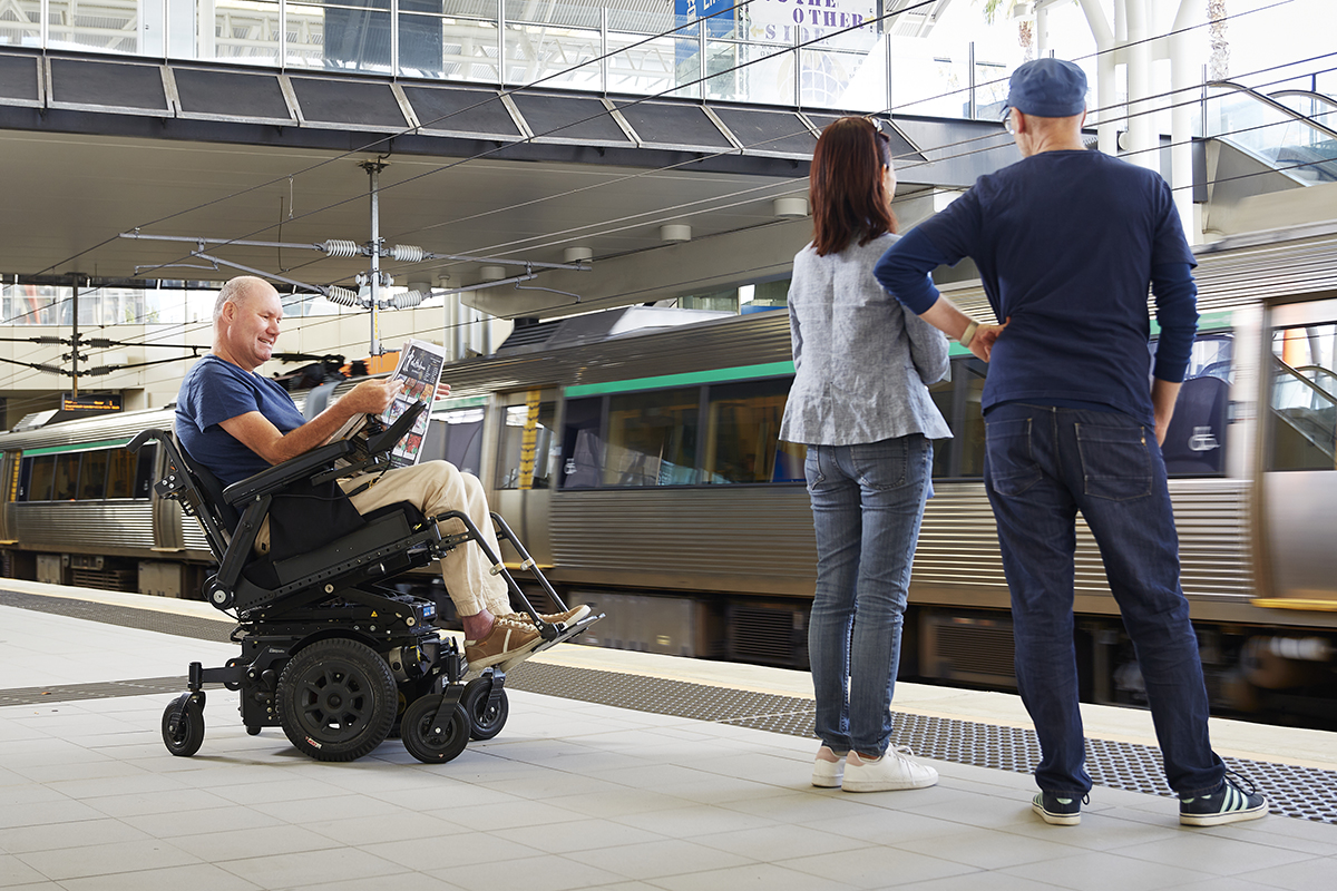 Man using power wheelchair at a train station, seating in tilt position while reading a magazine