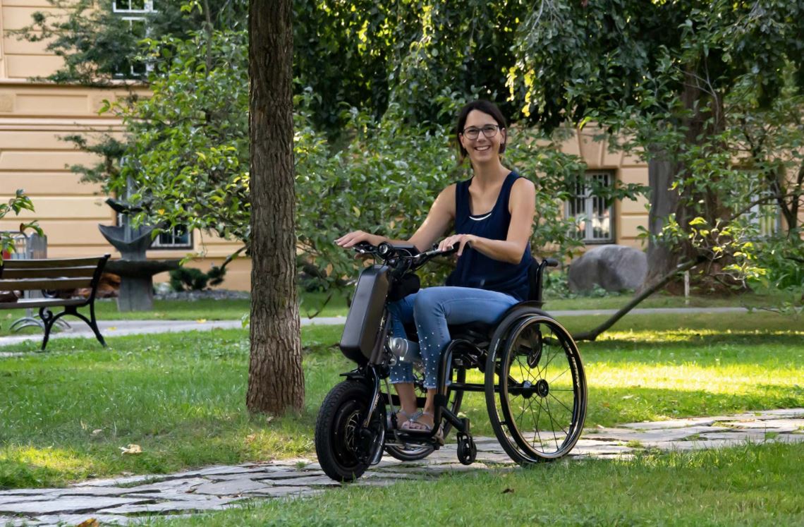 Manual wheelchair with Klaxon Race power assist attachment being used outdoors in a park setting