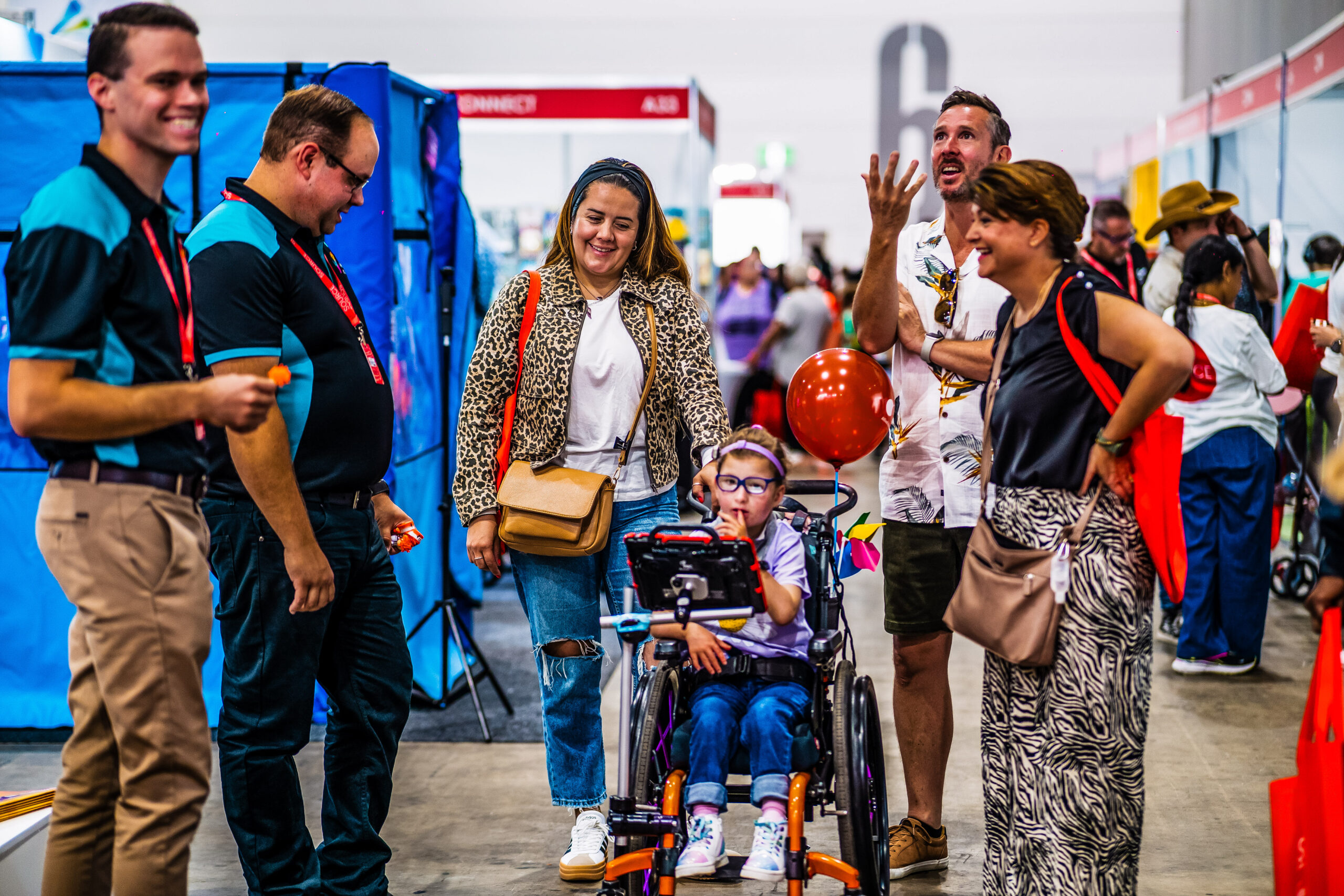 family and exhibitors interacting with child in wheelchair at disability expo showcasing assistive technology and mobility solutions